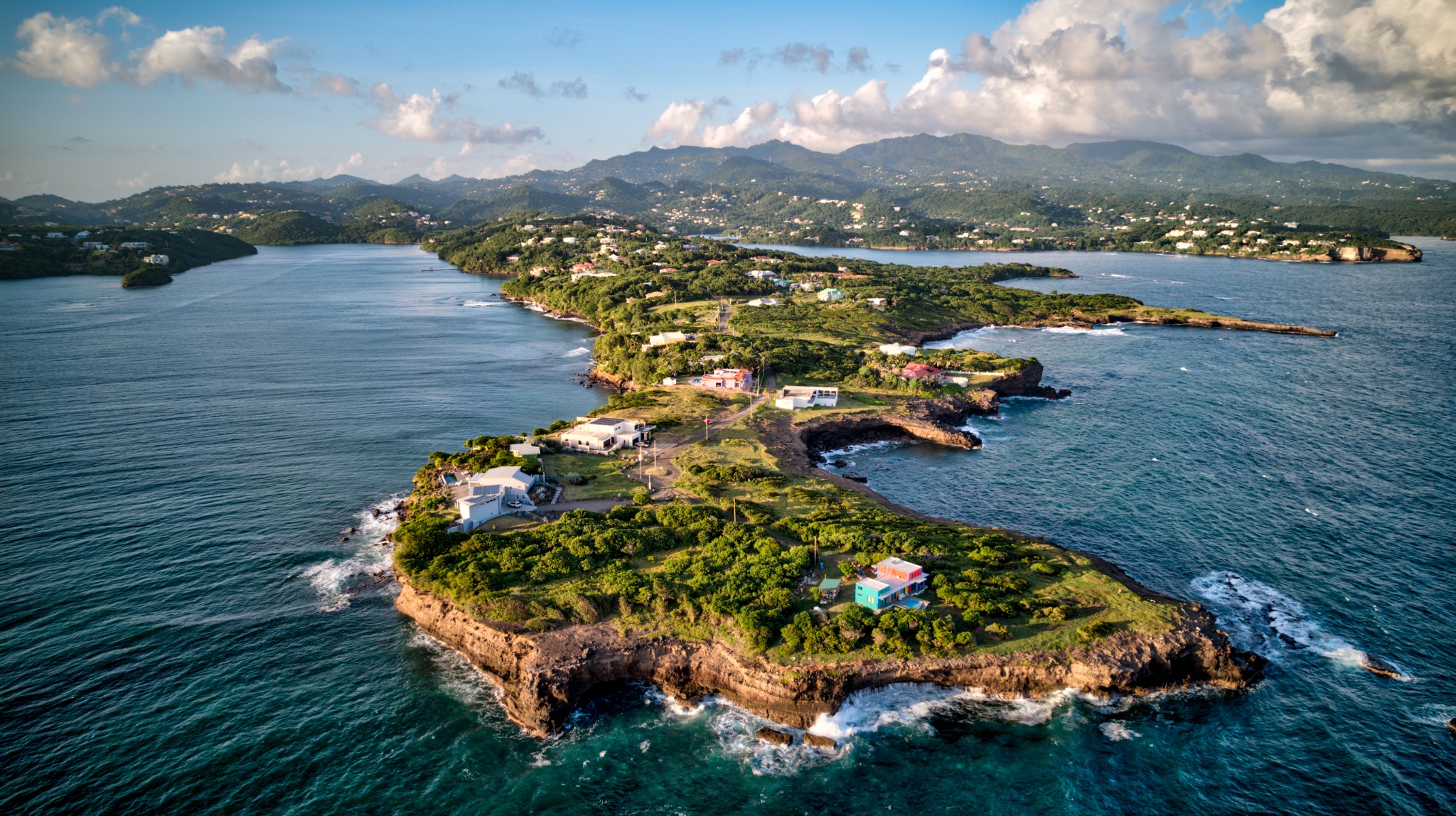 Aerial view of a wooded Caribbean peninsula surrounded by deep blue water, with hills behind